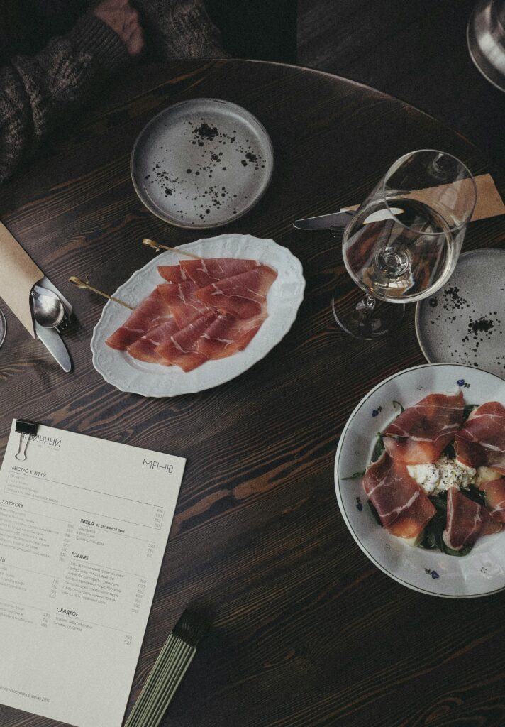 Overhead view of a dining table with prosciutto, wine, and menu.